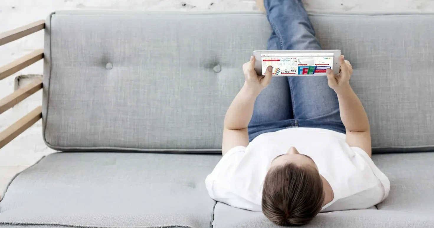 Eine Frau liegt mit den Beinen nach oben auf einem grauen Sofa, in der Hand hält sie ein Tablet, auf dem der THEORG-Onlineshop abgebildet ist.