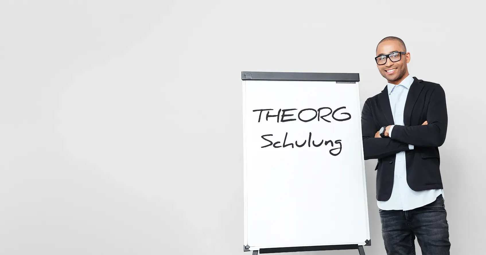 Ein junger Mann in Jeans, Hemd und Sakko lächelt in die Kamera, neben ihm steht ein Flipchart auf dem mit einer handschriftähnlichen Font "THEORG Schulung" steht.