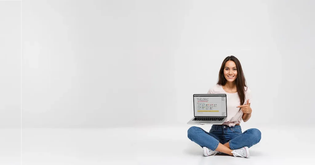 Eine junge lächelnde Frau sitzt im Schneidersitz vor einem weißen Hintergrund, in der rechten Hand hält sie einen Laptop, auf dem der THEORG-Startbildschirm abgebildet ist, sie zeigt mit der linken Hand darauf.